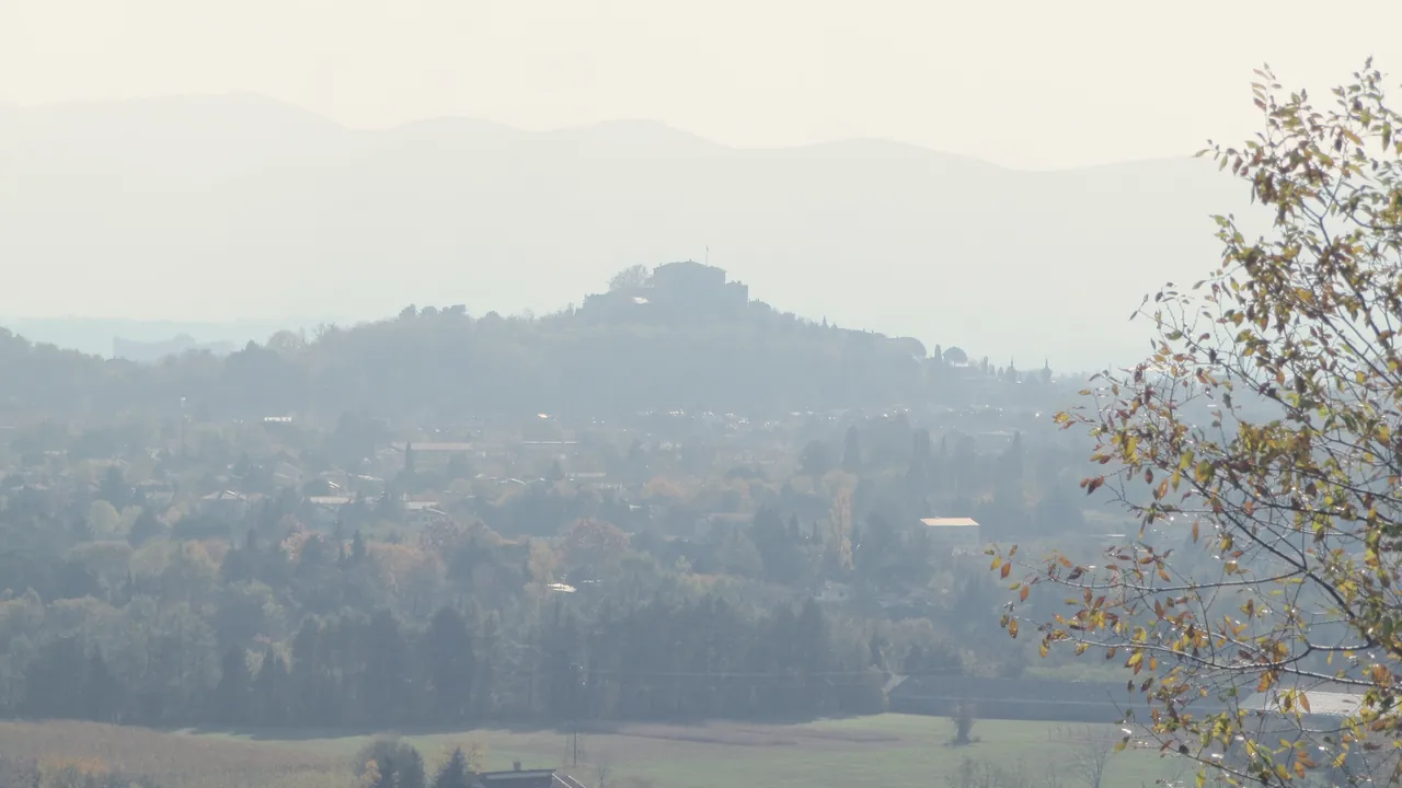 Hills veiled in autumn haze towards Gorizia, the castle far away on a hill and a yellow tree on the right.