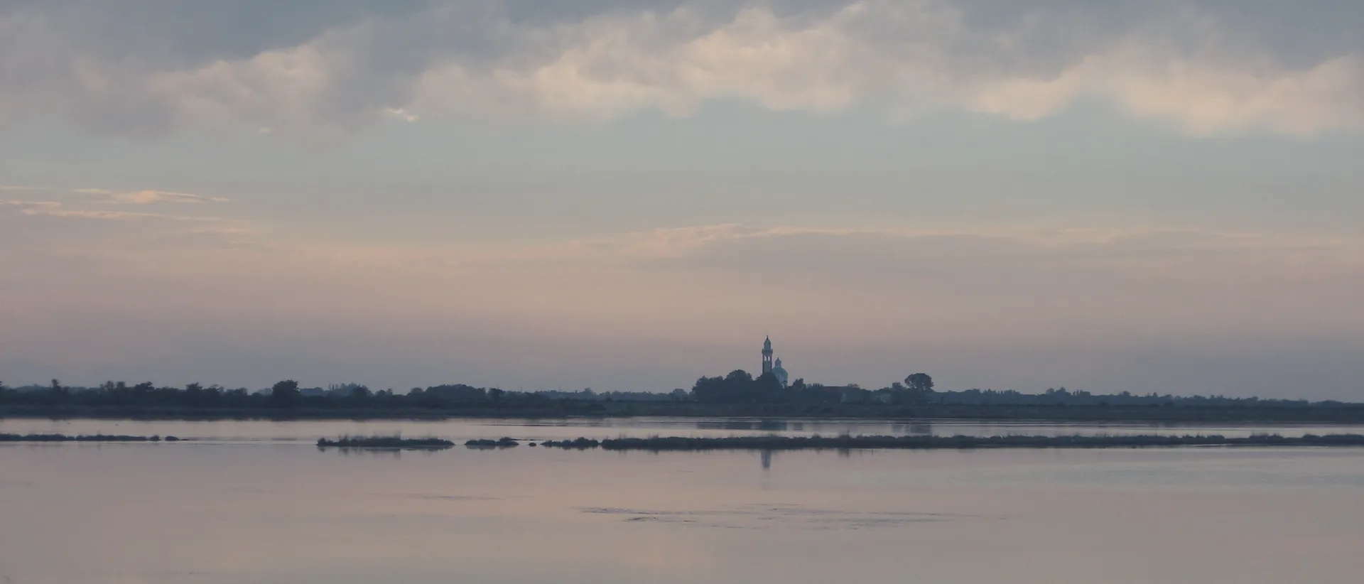 The Grado lagoon at sunset: still water and a pink sky, the island of Barbana with its bell tower on the horizon.