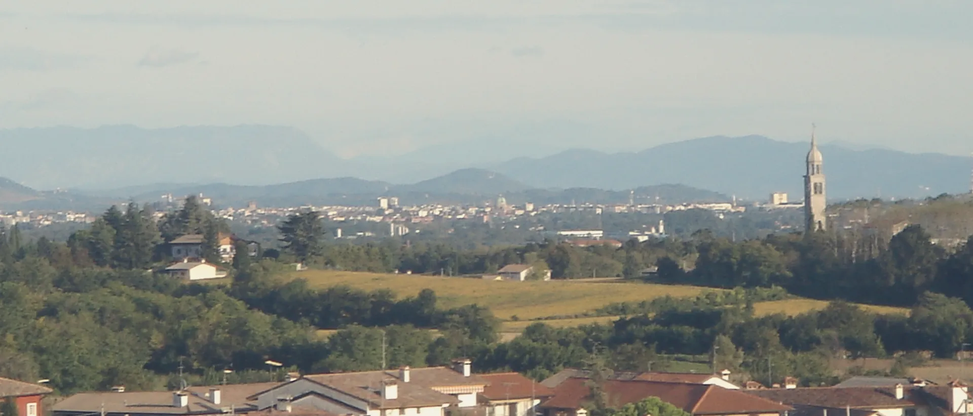The Friulian plain towards Udine seen from the hills, the city far away under a wide sky.