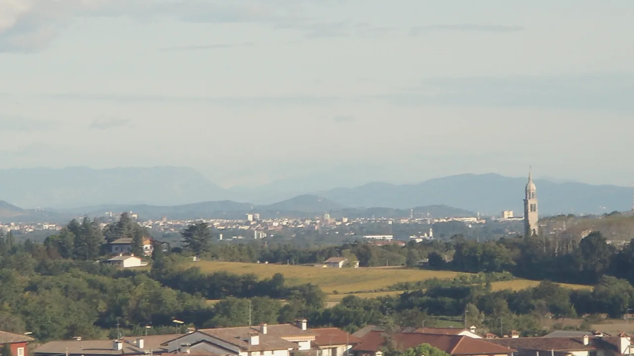 The Friulian plain towards Udine seen from the hills, the city far away under a wide sky.
