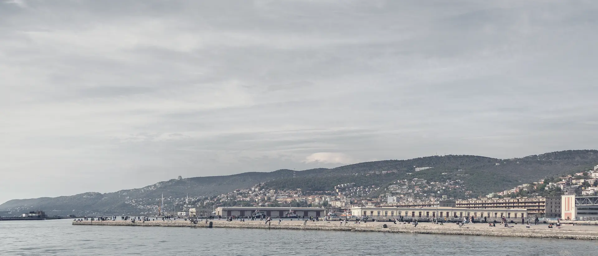 Trieste seen from the sea, with the pier in the foreground and the city under a wide sky.