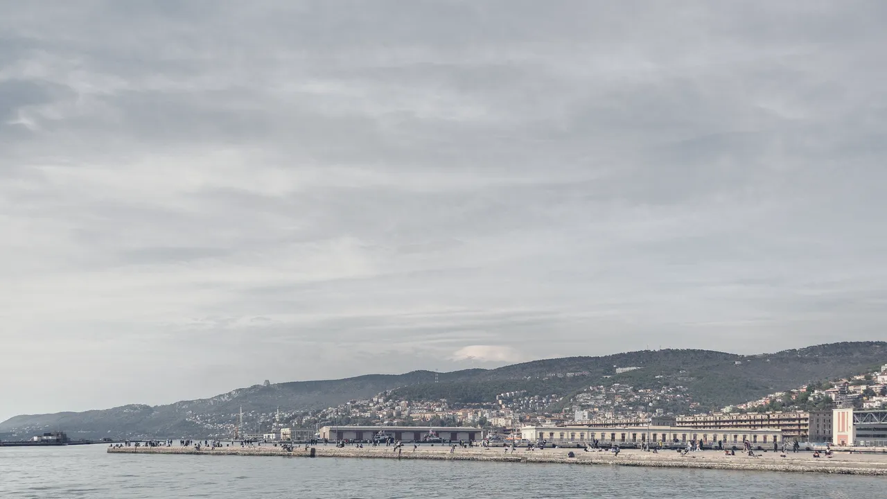 Trieste seen from the sea, with the pier in the foreground and the city under a wide sky.